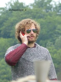 a man with curly hair and glasses talking on a cell phone while standing in front of trees