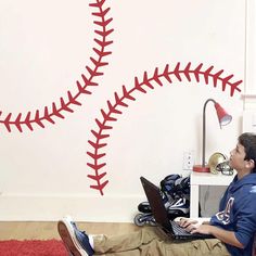 a man sitting on the floor using a laptop computer with a baseball wall decal behind him