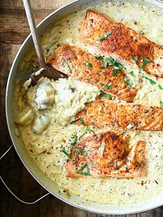 a skillet filled with salmon and vegetables on top of a wooden table next to a spoon