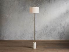 a floor lamp with a white shade on it in front of a concrete wall and wooden floor