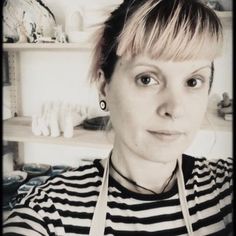 a woman with short hair wearing an apron and looking at the camera while standing in front of shelves