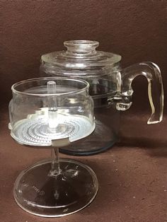 a glass tea pot with a clear lid on a brown tableclothed surface next to an empty cup and saucer