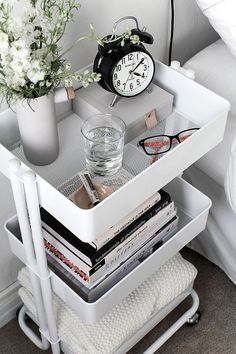 a white nightstand with magazines, glasses and a clock on the shelf next to it