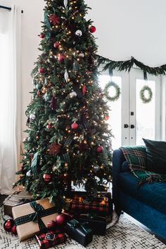 a christmas tree with presents under it in front of a blue couch and white curtains