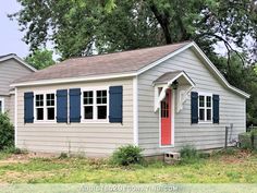 a gray house with blue shutters and a red door in the grass next to some trees