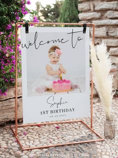 a welcome sign with a baby's first birthday cake on it and flowers in the background