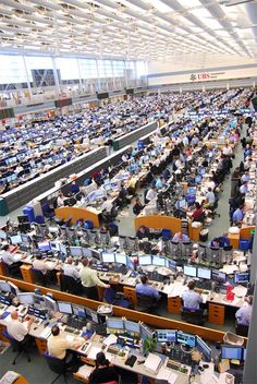 an office filled with lots of computers and people working at desks in front of large windows