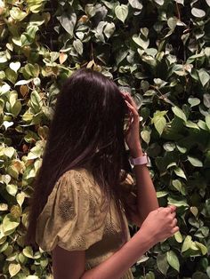 a woman standing in front of a bush with her hand on her head and looking at the ground