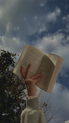 a person holding an open book in front of a blue sky with clouds and trees