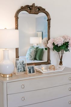 a white dresser topped with a mirror and vase filled with pink flowers next to a lamp