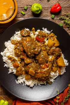 a black plate topped with rice covered in meat and veggies next to a glass of orange juice