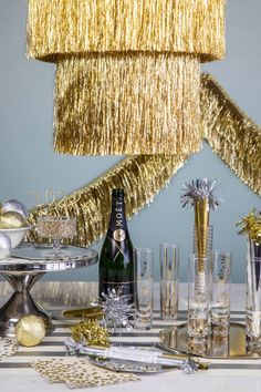 A table with a champagne bottle and glasses, adorned with Meri Meri's Gold Tinsel Chandelier. Tinsel Colorful Lights, White And Gold Party New Ywars, New Years Celebration Gold, Luxury Festival Celebration Danglers, Carnival New Years Party, Metallic New Years Party, Tinsel Chandelier, Gold New Years Party, Holiday Event Decor