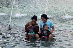 three women and two children are sitting in the water near a fountain that is spouting water