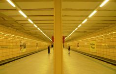 two people are standing in the middle of an empty subway station with yellow tiles on the walls