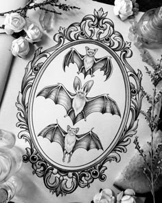 a black and white drawing of two bats in a frame surrounded by flowers on a table