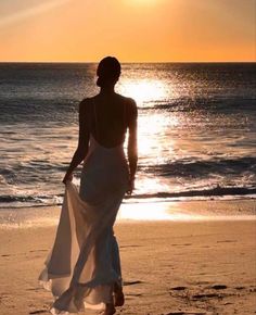 a woman walking on the beach at sunset with her dress blowing in the wind as the sun sets