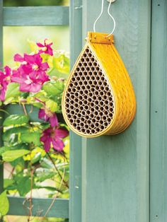 a yellow basket hanging on the side of a green fence next to pink flowers and greenery