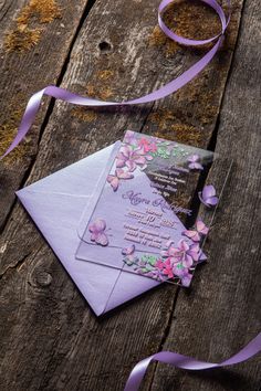 a purple wedding card on top of a wooden table next to a ribbon and some flowers