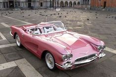 an old pink convertible car parked in front of a building with pigeons on the ground