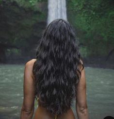 a woman standing in front of a waterfall with her back turned to the camera and long dark hair