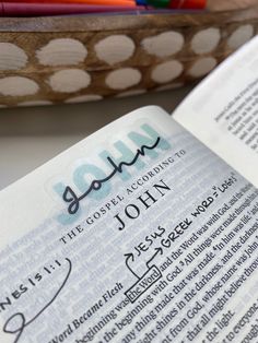 an open bible with the word john written in cursive writing on it next to a bowl of pencils