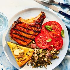 a white plate topped with grilled meat, tomatoes and beans next to a glass of water