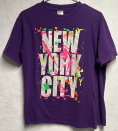 Purple tee shirt features short sleeves and New York City graphic with neon splatter accents. This item is old and it shows some signs of wear. The graphic is crackled and there is some pilling and discoloration in the underarm area (see photos). This item is listed as an adult M and is in good vintage condition with no holes. Urban Purple Short Sleeve T-shirt, Purple Grunge T-shirt With Letter Print, Multicolor Letter Print Grunge T-shirt, Multicolor Grunge T-shirt With Letter Print, Casual Paint Splatter T-shirt For Streetwear, Short Sleeve Graphic Tee With Paint Splatter, Casual T-shirt With Paint Splatter For Streetwear, Graphic Tee With Paint Splatter And Crew Neck, Paint Splatter Graphic Tee With Crew Neck