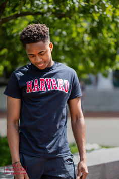 a young man wearing a harvard t - shirt looking at his cell phone while standing in front of a tree