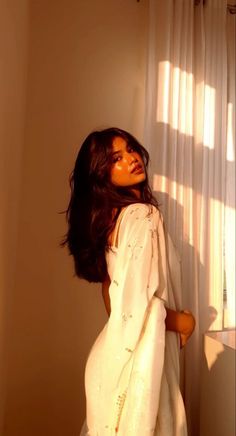 a woman standing in front of a window wearing a white dress and holding her arms behind her back