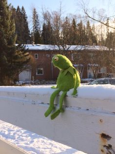 a green stuffed animal sitting on top of a white fence next to snow covered ground