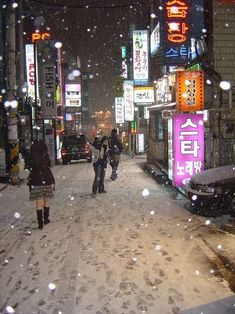 people are walking in the snow on a city street at night with neon signs and cars