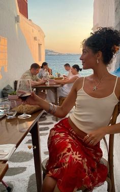 a woman sitting at a table in front of the ocean with people eating and drinking