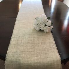 a table runner with white flowers on it and an empty vase in the middle, sitting on a dining room table