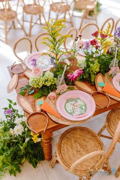 the table is set with pink plates and flowers on it, along with wicker chairs