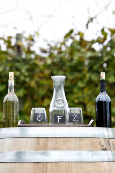 three wine bottles and two empty glasses sitting on top of a wooden barrel with trees in the background