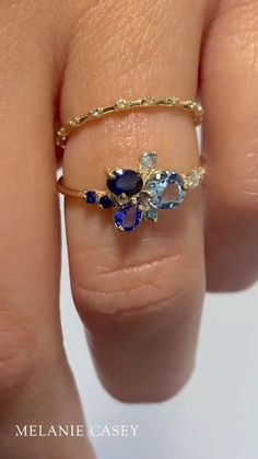 A blue gemstone cluster ring is worn by a woman's hand. Luxury Blue Topaz Ring With Rose Cut Diamonds, Blue Cluster Rings With Multi-stone, Blue Topaz Ring With Rose Cut Diamonds For Anniversary, Anniversary Blue Topaz Ring With Rose Cut Diamonds, Blue Multi-stone Cluster Ring, Blue Oval Multi-stone Cluster Ring, Blue Cluster Rings With Accent Stones, Blue Rings With Rose Cut Diamonds For Promise, Blue Rose Cut Diamond Promise Ring