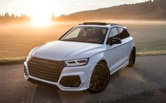 the front end of a white car parked on a road near grass and trees at sunset