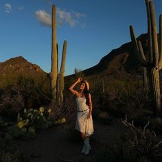 a woman in a white dress standing next to some cacti on a dirt road