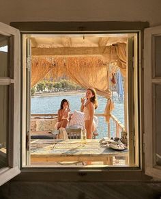 two women sitting on a bed in front of an open window looking at the water