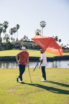 two men walking in the grass with an umbrella over their heads and one holding a golf club