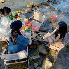 three women sitting at a table by the water eating food and drinking beer while another woman looks on