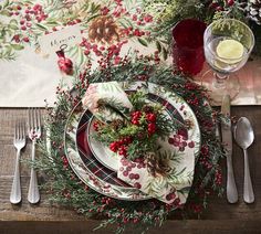 a place setting with holly, berries and pine cones on the table for christmas dinner