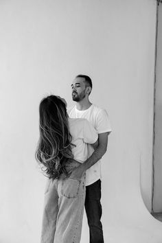 a man and woman standing next to each other in front of a white wall with their arms around each other