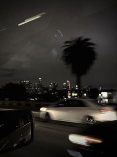 cars driving down the road at night with city lights in the background and palm trees