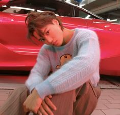 a young man sitting on the ground next to a red car