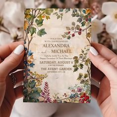 two hands holding up a wedding card in front of flowers and greenery on the table