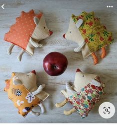 four stuffed animals are arranged around an apple on a table with other fabric toys in the shape of hedges