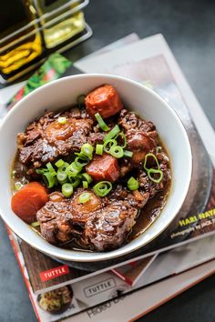 a white bowl filled with meat and vegetables on top of a table next to magazines