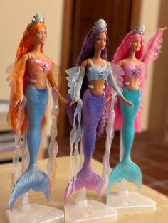 three barbie dolls are standing on the counter top, one is wearing a mermaid costume