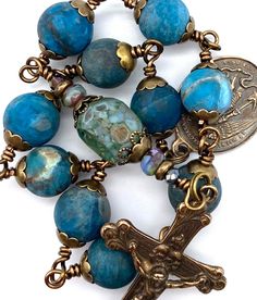 "Beautiful handcrafted vintage-inspired Catholic Heirloom Tenner Rosary with large Pacific Blue Apatite Matte gemstones. Pater bead is an Aquamarine Czech 2-way cut glass bead with Czech glass accent beads and Antique Brass bead caps. Each bead is wire wrapped using 20-gauge Vintage Bronze colored wire for strength and durability. The intricate design of the crucifix and medal are antique-styled bronze replicas giving the Rosary that \"Old World\" finish. Specifics: *Ave Beads: Pacific Blue Apat Bohemian Hand-strung Blue Beads, Artisan Blue Beads For Jewelry Making, Handmade Artisan Blue Beads, Gems, And Cabochons, Bead Wire, The Rosary, Pacific Blue, Blue Apatite, Bead Caps, Bronze Color